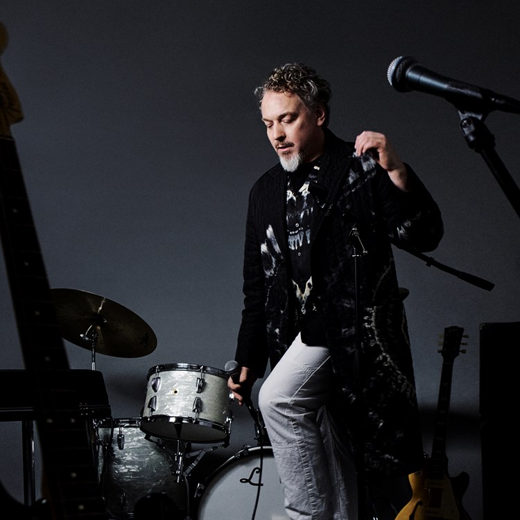 Bild på artisten Eric Gadd stående mitt bland olika musikinstrument mot en mö...