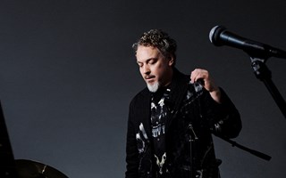 Bild på artisten Eric Gadd stående mitt bland olika musikinstrument mot en mö...