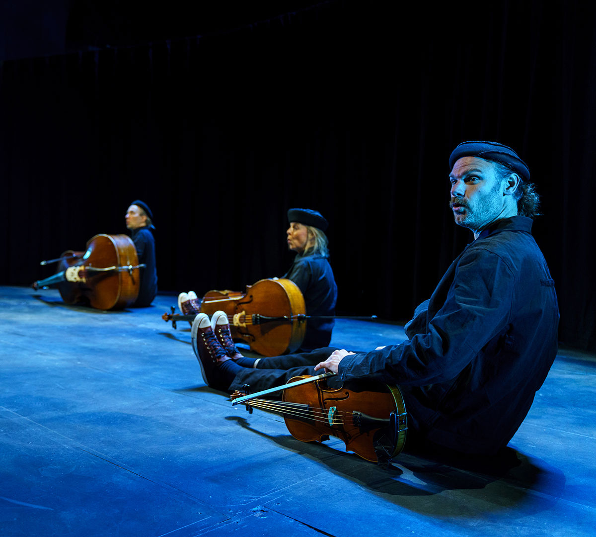 Bild på musikerna i konserten Familjen Stråke sittandes på rad på golvet i mörka kläder och små mössor. Framför sig har de sina stråkinstrument. 