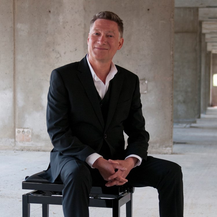 Helbild av pianisten Mårten Landström sittande på en pianopall i en betongmiljö.