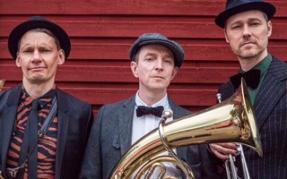 Musikerna i Brass Deluxe står uppradade vid en röd träfasad