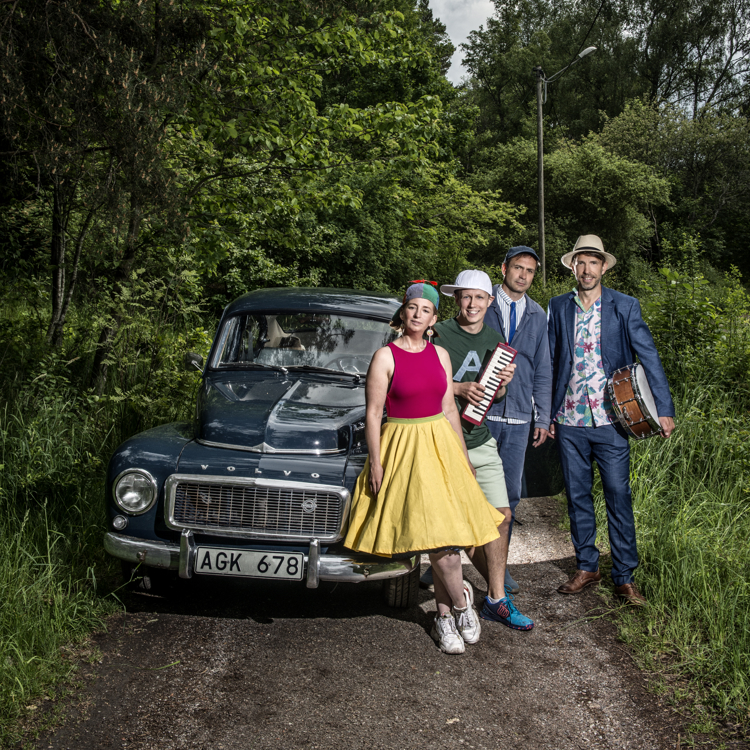 De medverkande i konserten Familjen Svängsson står utomhus, lutande mot en gammal Volvo-bil, iklädda fina kläder. Några av medlemmarna håller i musikinstrument.