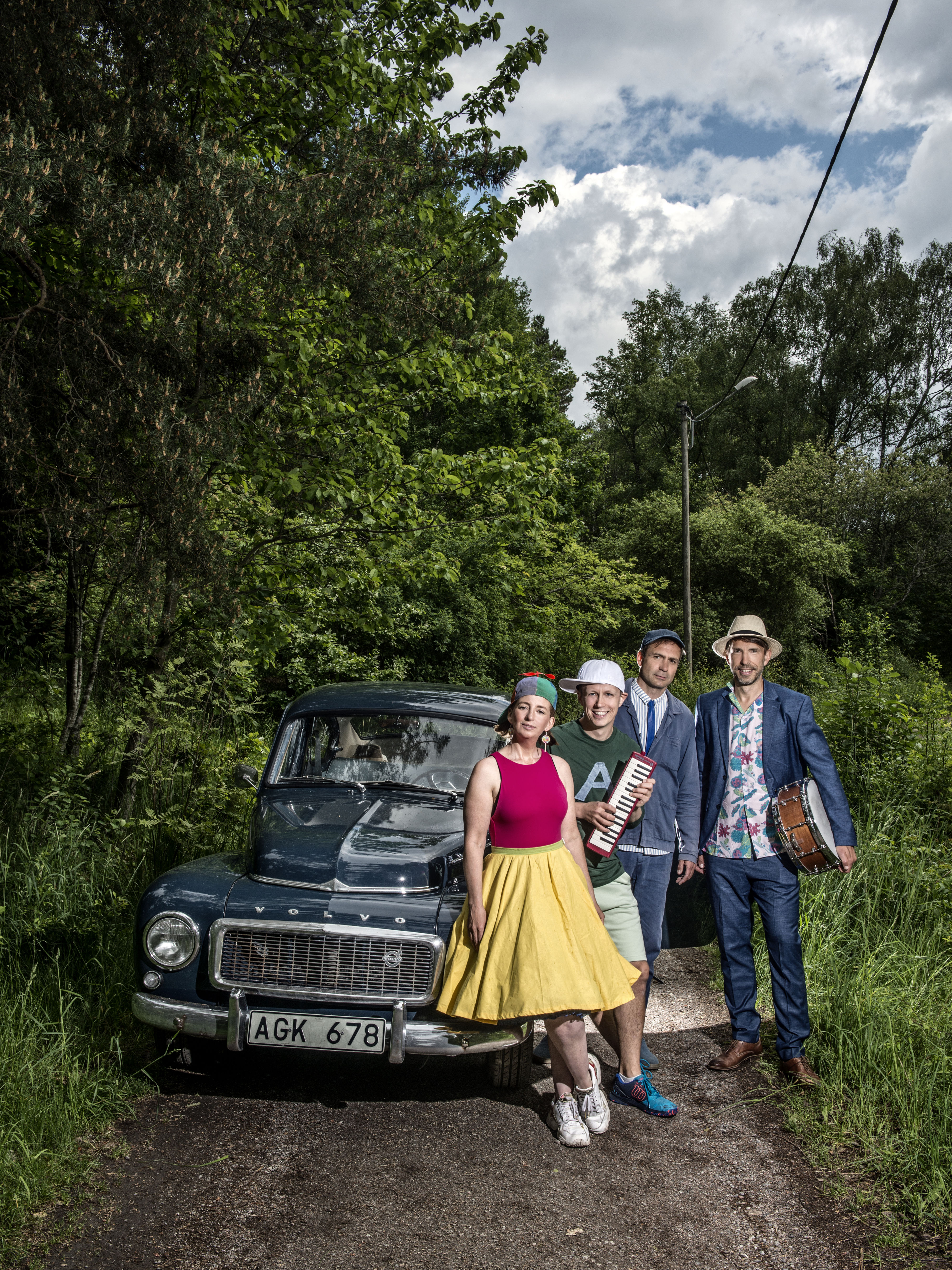 De medverkande i konserten Familjen Svängsson står utomhus, lutande mot en gammal Volvo-bil, iklädda fina kläder. Några av medlemmarna håller i musikinstrument.