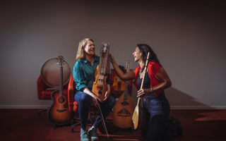 Multimusikerna Emilia Amper och Nadin Al Khalid sitter på ett trägolv. Runt om dem står en massa olika musikinstrument. Ansiktsuttrycken utstrålar glädje.