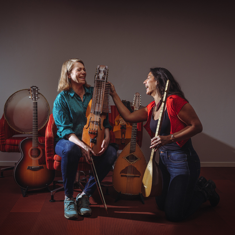 Multimusikerna Emilia Amper och Nadin Al Khalid sitter på ett trägolv. Runt om dem står en massa olika musikinstrument. Ansiktsuttrycken utstrålar glädje.