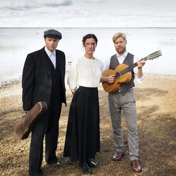 De tre musikerna och artisterna i föreställningen Swede Hollow står i tidsenliga kläder vid en strandkant. Bakom dem är det vatten. Deras ansiktsuttryck är allvarliga och ledsna.