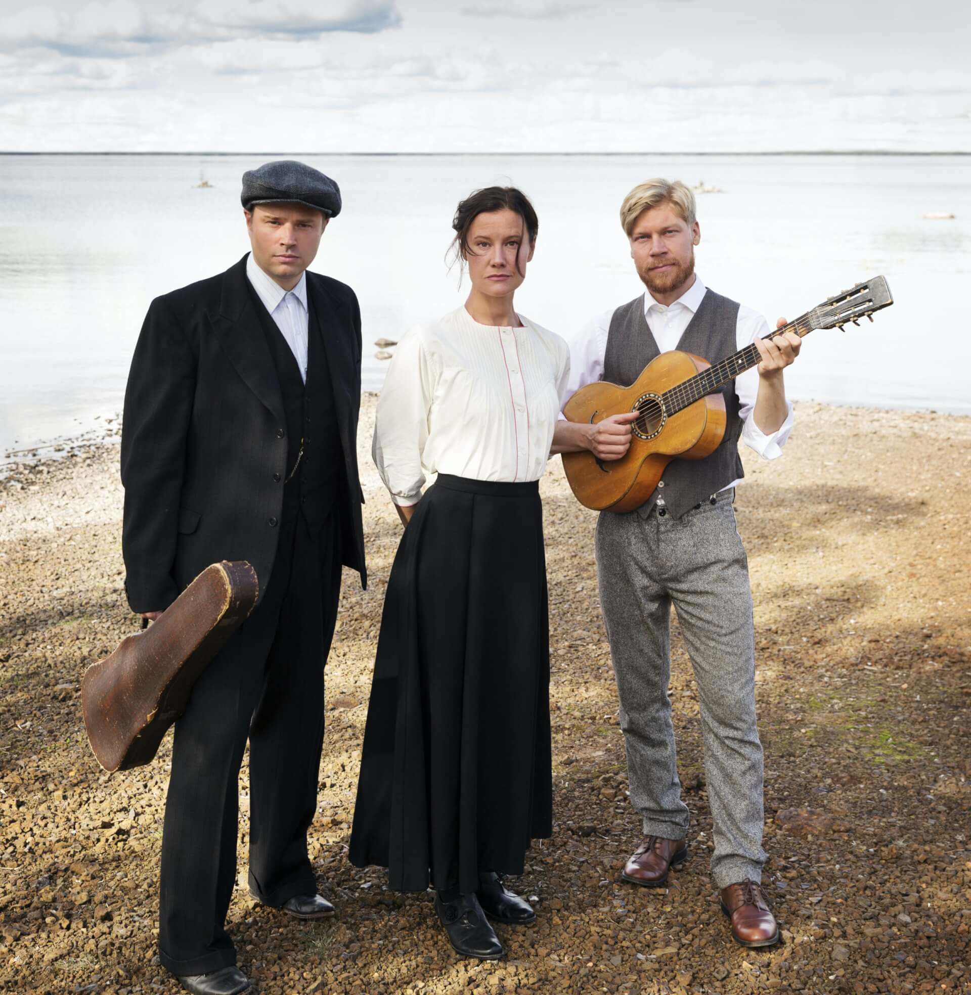 De tre musikerna och artisterna i föreställningen Swede Hollow står i tidsenliga kläder vid en strandkant. Bakom dem är det vatten. Deras ansiktsuttryck är allvarliga och ledsna.