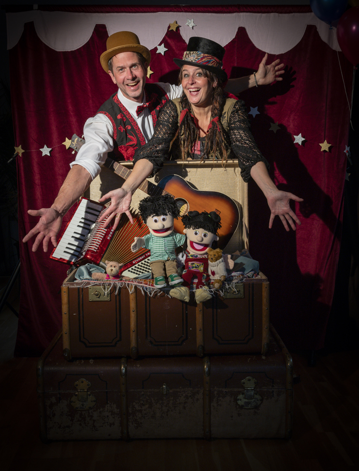 En glad kvinna och man sträcker ut händerna och visar upp en väska med instrument, dockor och en nalle.