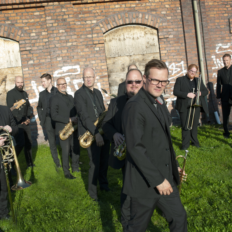 Ensemblebild av Norrbotten Big Band. Orkesterns musiker står i kostym, utspridda framför en tegelvägg.