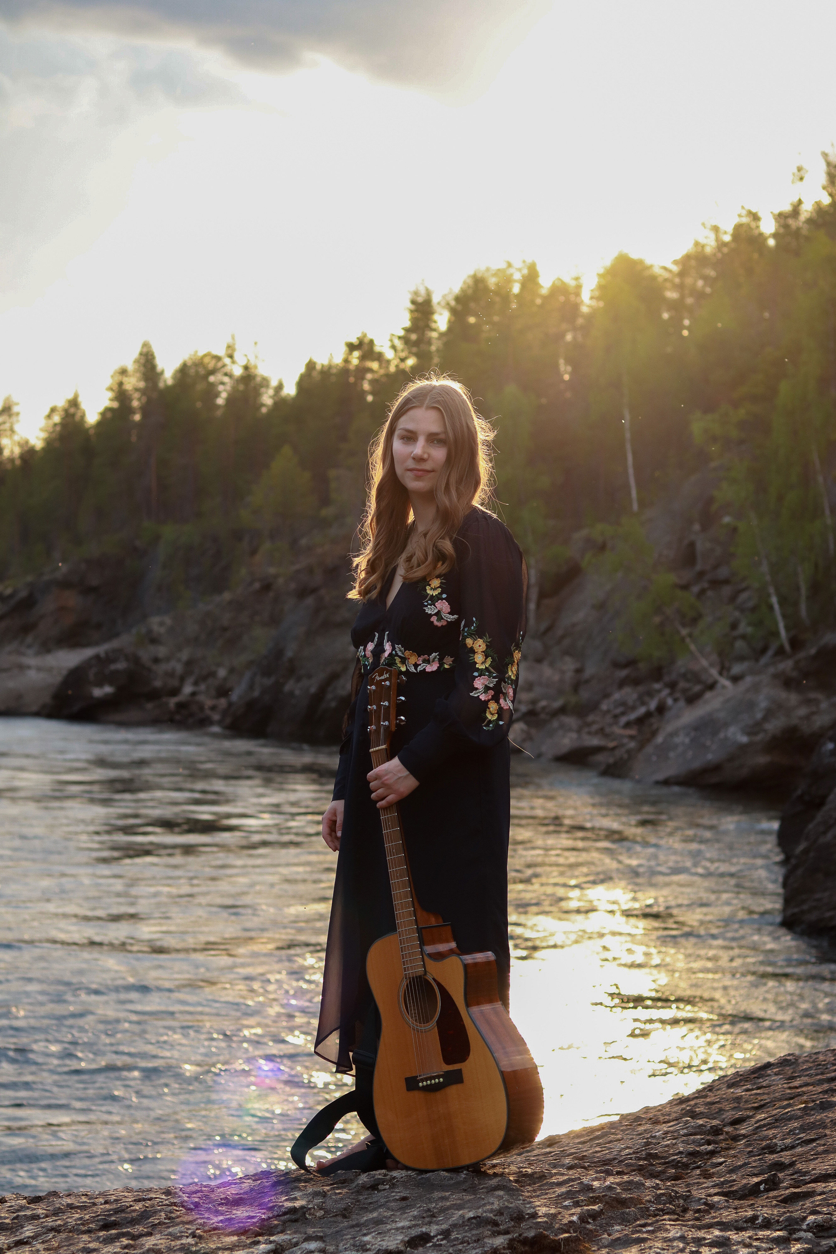 Bild på musikern Klara Pasma stående på en sten vid en fors. I handen håller hen en gitarr.