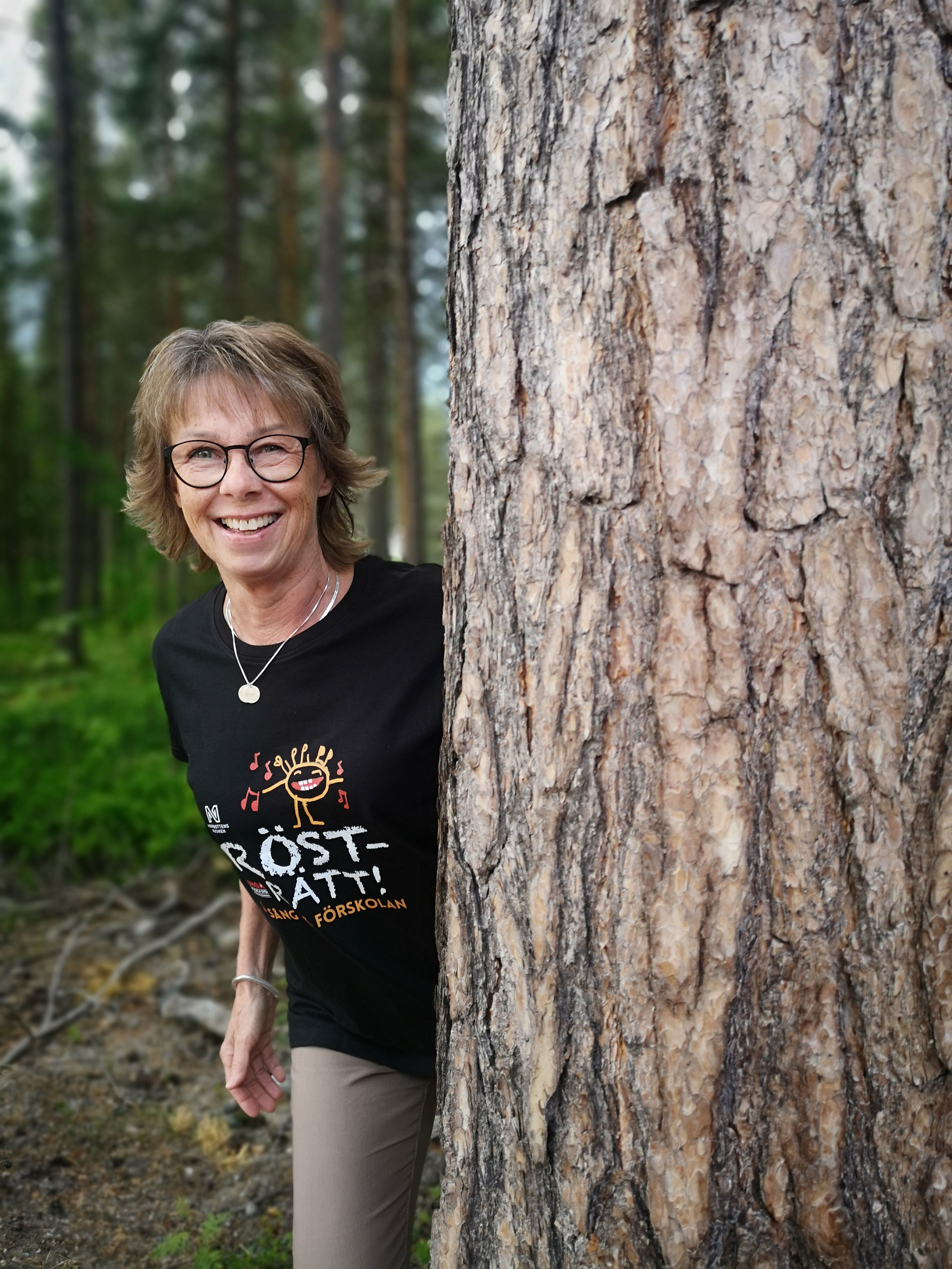 Rösträttscoach Carina Higberg tittar fram bakom en trädstam. Bakom henne ser men skog.