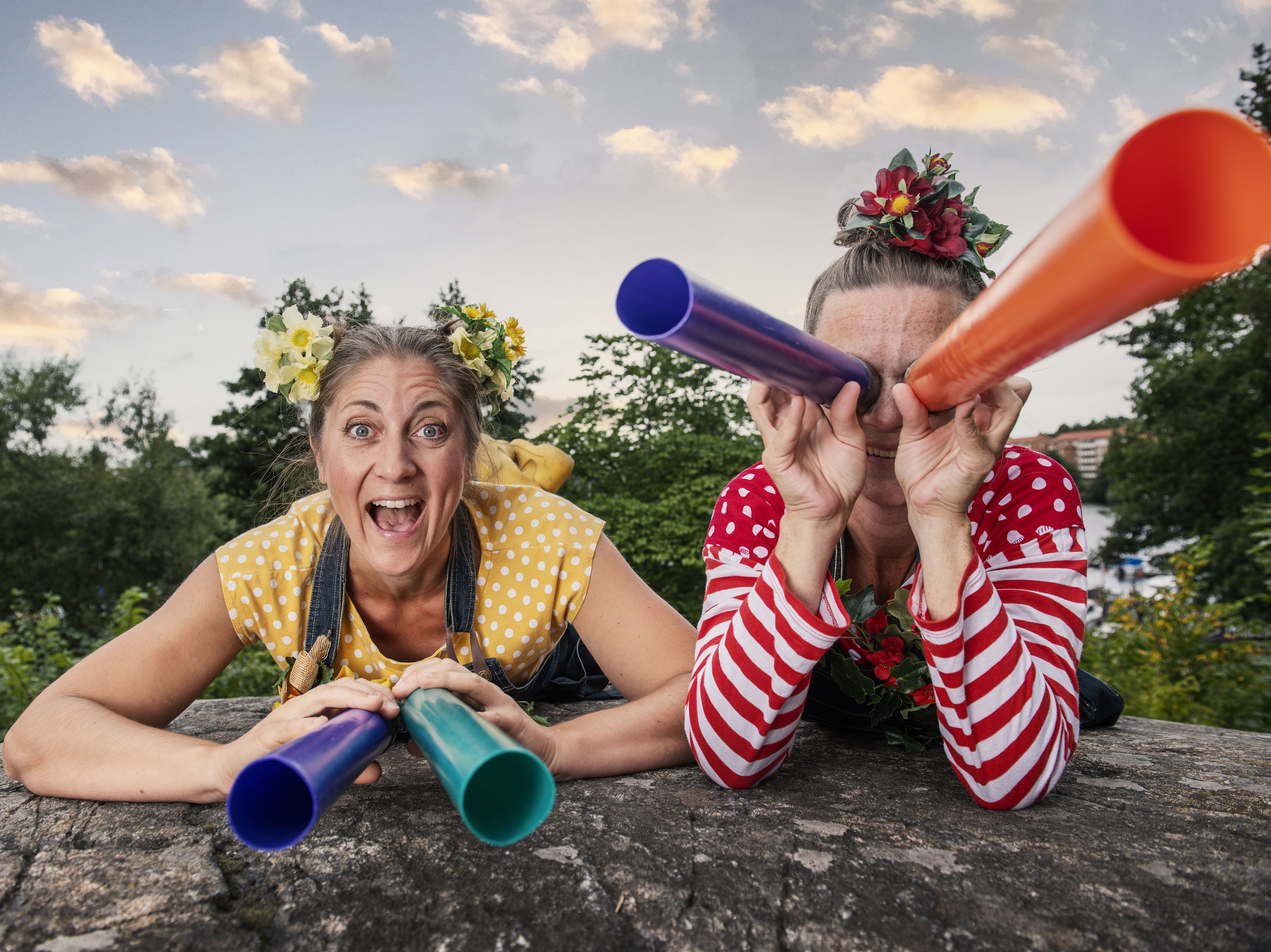 Två färgglada kvinnor ligger och spanar - den ena med kikare för ögonen, den andra med kikare på marken.
