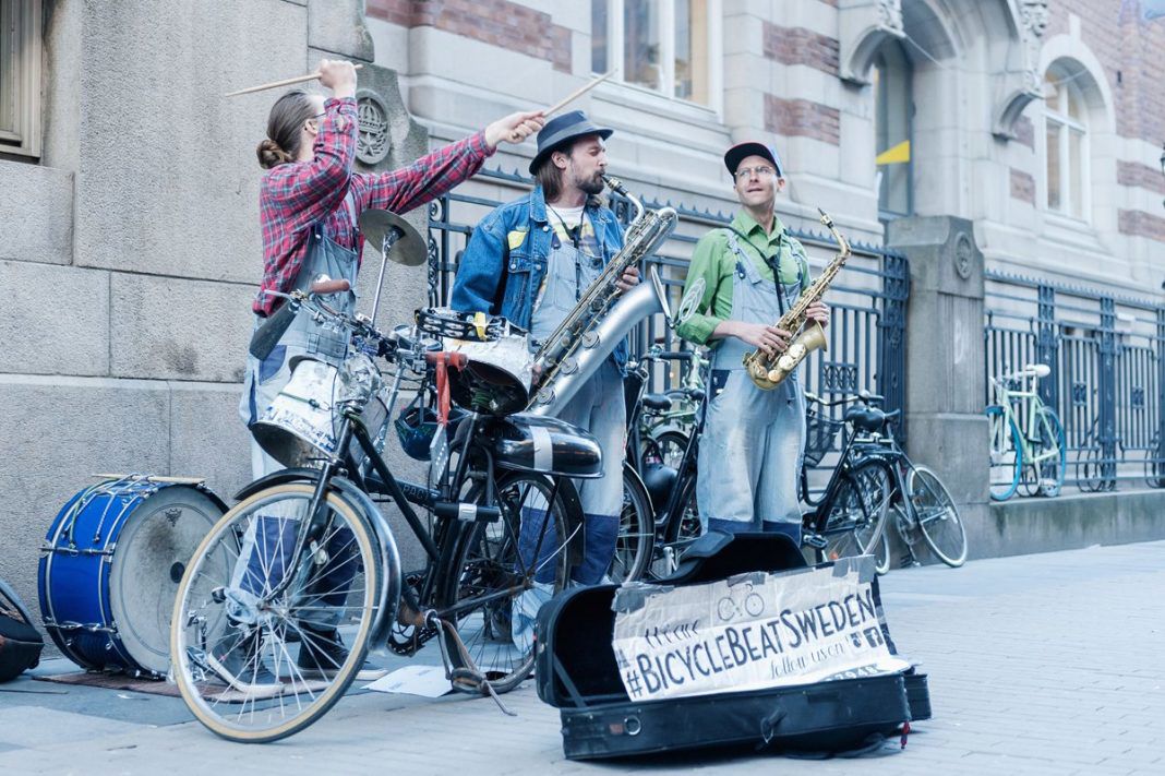 Bild på gruppen Bicycle Beat. De tre musikerna står ute på gatan. I händerna och framför sig har de sina musikinstrument. Det är allt från en saxofon till en cykel. 