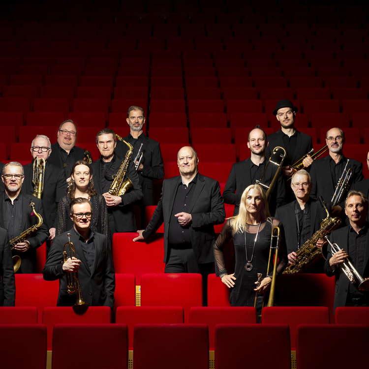 Ensemblebild på musikerna i Norrbotten Big Band stående i mörka kostymer bland stolsrader i ett konserthus. I mitten står bandets konstnärliga ledare Joakim Milder. 