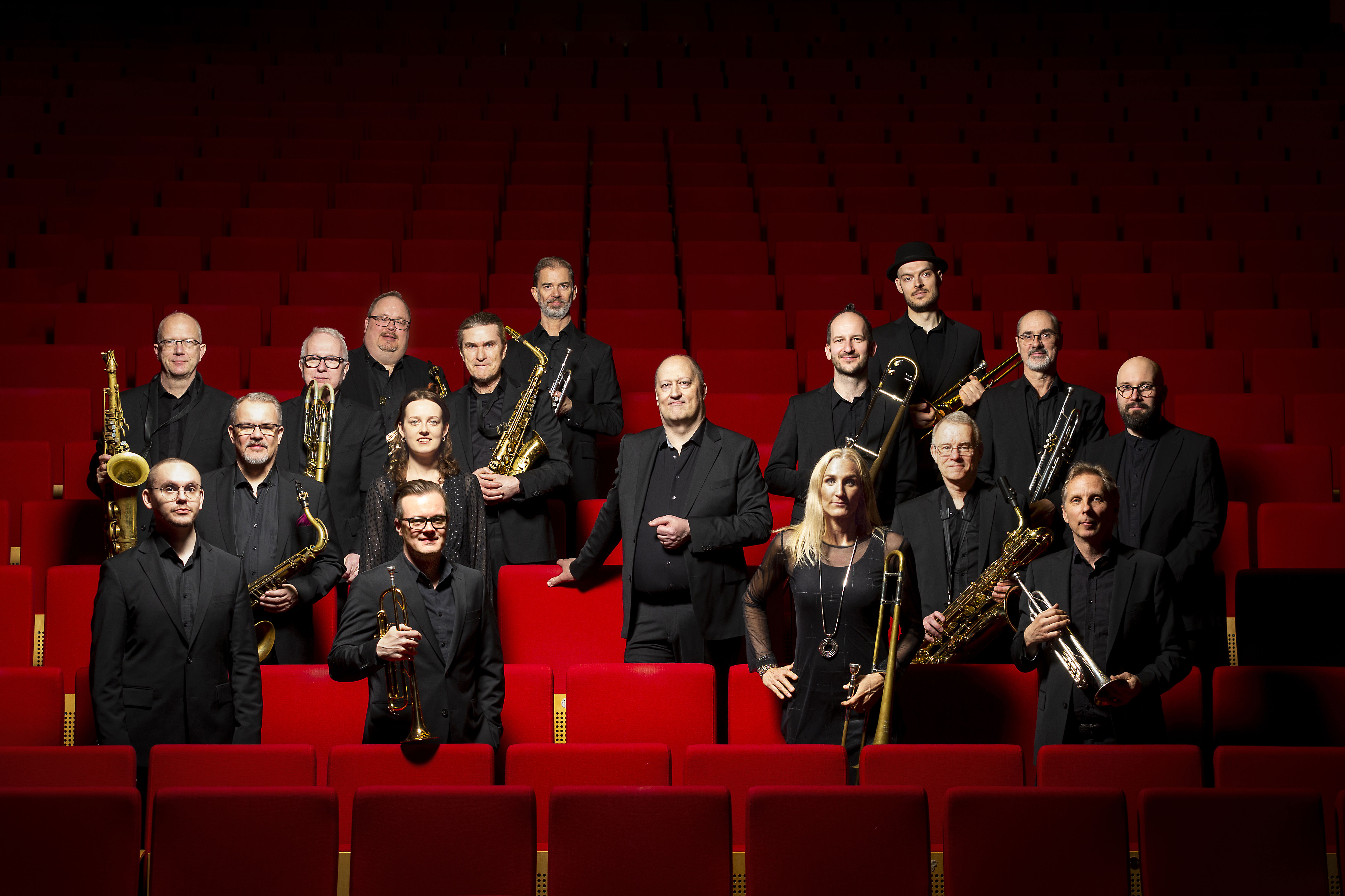 Ensemblebild på musikerna i Norrbotten Big Band stående i mörka kostymer bland stolsrader i ett konserthus. I mitten står bandets konstnärliga ledare Joakim Milder. 